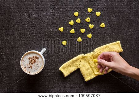 Cup of coffee, yellow knitted sweater for a doll and a heart-shaped buttons on a grunge black background. Female hand holding a button.