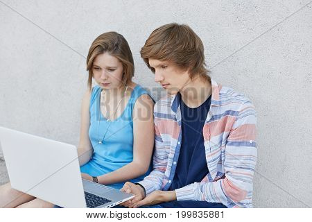 Stylish Teenagers Sitting Next To Each Other, Looking Attentively In Screen Of Laptop, Reading News
