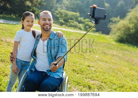 Collecting memories. Pleasant young man with disabilities hugging his adorable little daughter and taking a selfie with her, using a selfie stick