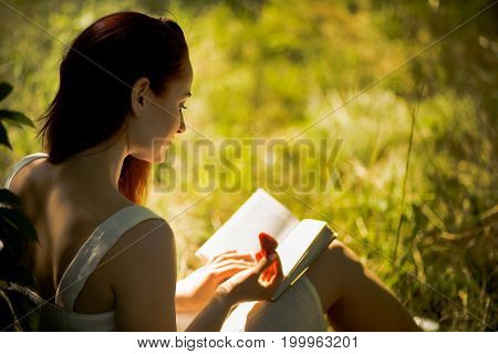 Rear view of attractive woman sitting on grass with book in her hands. Young foxy haired woman reading book in nature. Toned image.