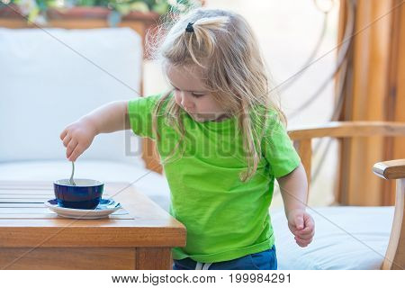 Little kid boy making tea. Childhood and baby care concept. Boy child mixing sugar in blue cup. Child boy with blond long hair mixing sugar with teaspoon. Healthy breakfast for child.