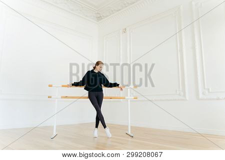 Classic Ballet Dancer Has Stretching Workout, Stands Near Ballet Barres, Wears Black Sweatshirt And 