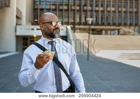Mature african businessman eating sandwich outdoor. Senior entrepreneur standing outside office building, looking away while enjoying vegetable sandwich. Business man having lunch on the move.