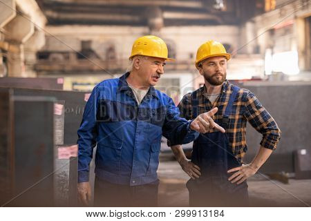 Aged confident master in hardhat and workwear pointing at one of industrial machines while showing it to his trainee