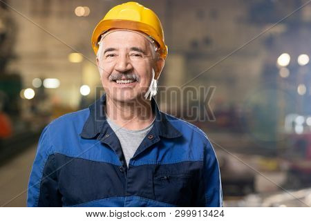 Senior cheerful professional in sphere of machinebuilding looking at you while standing inside large industrial workshop