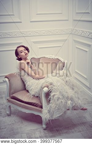 Portrait of a beautiful chic woman posing in a luxury white dress in vintage armchair. Evening makeup and hairstyle of the 20s. 