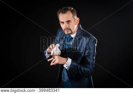 Handsome Male Magician With A Beard And Mustache In A Blue Tailcoat Holding A Dove On A Black Backgr