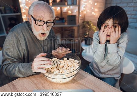Grandson And Grandfather Are Sitting At The Table And Watching A Movie With Popcorn. Grandson Closes