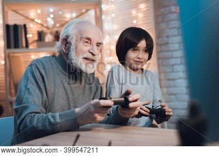 Grandfather And Grandson Are Playing Video Games On Computer At Night At Home.