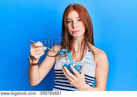 Young red head girl eating measure meter doing diet for weight loss relaxed with serious expression on face. simple and natural looking at the camera. 