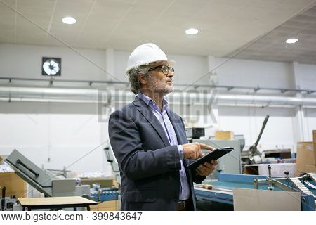 Serious Supervisor In White Helmet Holding Tablet And Looking On Manufacturing Process. Portrait Of 