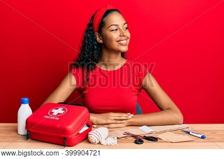 Young african american girl sitting on the table with first aid kit making cure looking to side, relax profile pose with natural face and confident smile. 