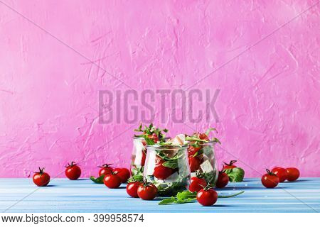 Salad With Arugula, Cheese, Cherry Tomatoes And Prosciutto In Glass Jars, Pink Blue Background, Sele