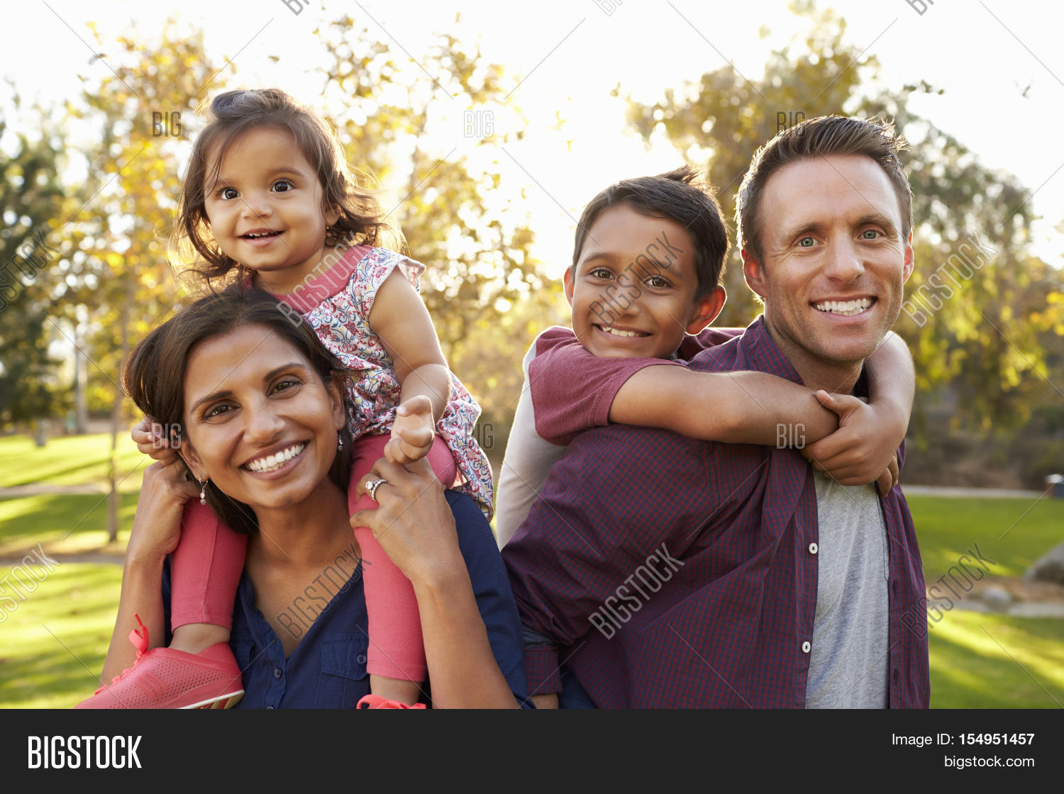 mixed race parents carry their kids piggyback in a park
