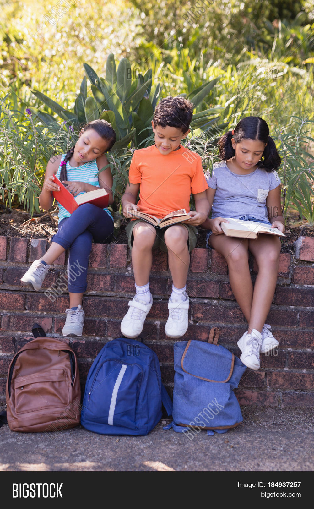 smiling friends reading books while sitting on retaining wall at
